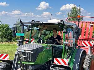 Auf dem Foto ist eine Frau zu sehen, die in einem grossen Traktor mit geschlossener Fahrerkabine sitzt.