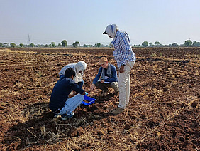 Five men work together on a plowed dry field and analyze soil samples with a blue bowl.