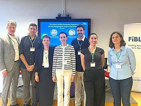 The seven participants of the roundtable discussion stand together in front of a screen. Four of them are women and three are men.