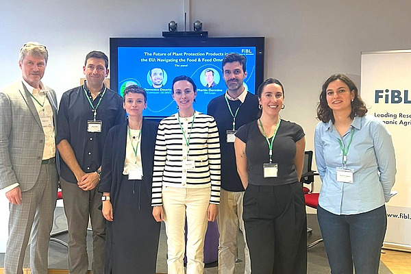 The seven participants of the roundtable discussion stand together in front of a screen. Four of them are women and three are men.