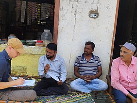 Four men, including a foreign researcher, sit in front of a house and conduct an interview with notes and paper.