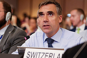 A man in a hall with a table microphone and a sign saying Switzerland.