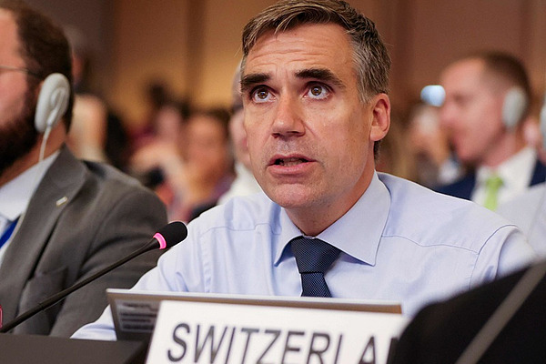 A man in a hall with a table microphone and a sign saying Switzerland.