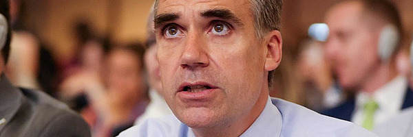 Ein Mann in einem Saal mit Tischmikrofon und Schild mit Switzerland drauf.