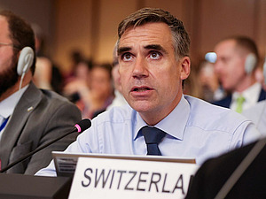 Ein Mann in einem Saal mit Tischmikrofon und Schild mit Switzerland drauf.