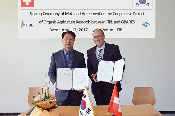 2 men stand at a table and present the signed agreements. THe table is decorated with a Swiss and a South Korean flag as well as some flowers.