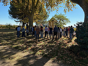 Gruppe unter Bäumen vor Gebäude.