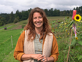 Eine Frau mit langen, gewellten, rötlich-braunen Haaren steht vor einem Garten und einer Kunstwiese. Im Hintergrund ist Wald und der Himmel ist bewölkt. Die Frau lacht freundlich und trägt einen orangenen Pullover und eine Weste aus Fell.