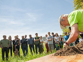 Mann, der etwas an einem Tisch mit Erde zeigt, im Vordergrund, Menschengruppe im Hintergrund. 