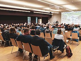 Teilnehmende in einem Konferenzsaal, die einer Rednerin zuhören.