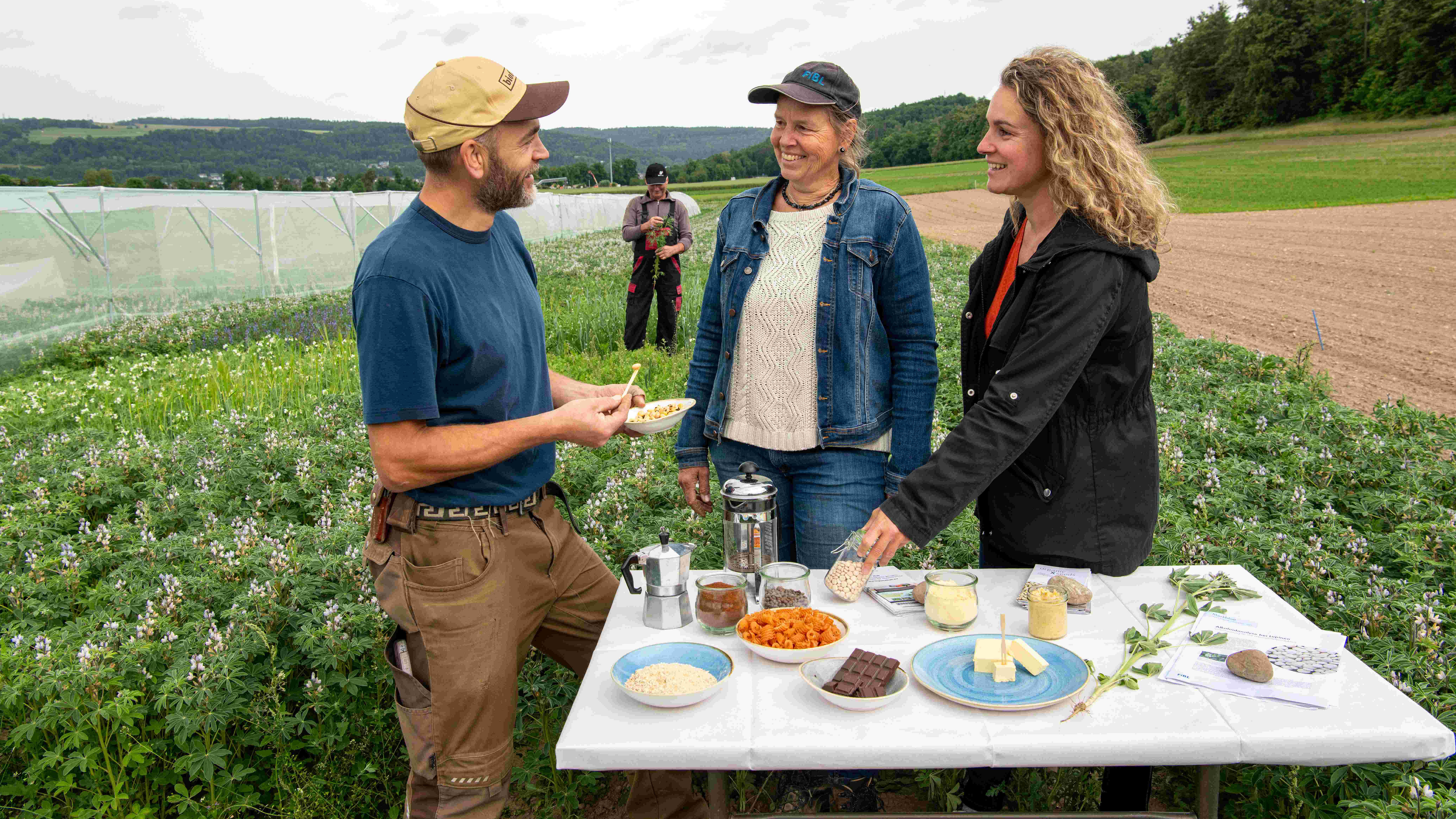 Ein Mann und zwei Frauen auf einem Feld an einem Tisch. 
