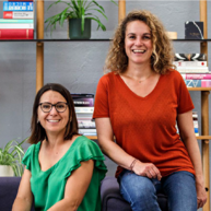 Zwei Frauen sitzen vor einem Holzregal mit Pflanzen und Büchern darin. Die eine trägt ein leuchtend grünes, die andere ein rotes T-shirt. Sie sind um die 30 Jahre alt und lächeln sehr offen und breit. Die eine sitzt etwas höher als die andere.