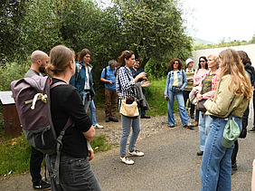 A group of people are standing in a circle on a path. In the middle, a woman is explaining something.