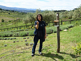 A woman is standing next to a wild bee hive. In the background, a varied landscape with herbs and olive trees can be seen.