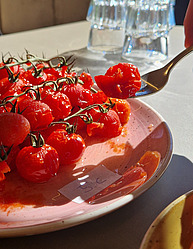 In einem Innenraum mit hohen Glasfenstern im Hintergrund sind leuchten rote, gekochte Tomaten auf einem Teller. Eine Gabel kommt von rechts ins Bild und nimmt eine der Tomaten.
