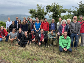 Gruppenfoto mit rund 18 Personen vor drei Bäumen und wenigen sichtbaren Reben