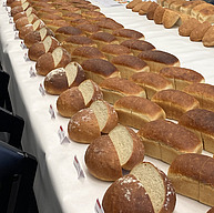 Auf einem Tisch mit einer weissen Tischdecke liegen vier Reihen mit runden und kastenförmigen, goldbraunen Broten. Die Brote in der ersten Reihe sind in der Mitte durchgeschnitten. Im Hintergrund ist ein Koch und eine weitere Reihe Brote zu sehen. Die Perspektive geht nach hinten. Die Brote sehen sehr lecker aus.