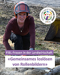 Eine Frau zwischen 30 und 40 kniet mit einer Motorsäge in der Hand und einem Bauhelm auf dem Kopf auf einer Wiese oder einem Acker. Sie schaut selbstbewusst in die Kamera und trägt ein Bordeauxrotes Oberteil und Arbeitshosen.